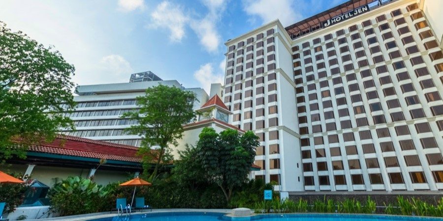 A hotel pool with clear water in front of a tall building, surrounded by lounge chairs and palm trees.